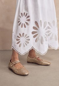 Auksiniai pinti bateliai su smailia nosimi ir dviem dirželiais. Batai turi reljefišką raštą ir žemą kulniuką. Fone matyti balta siuvinėta sijonė.