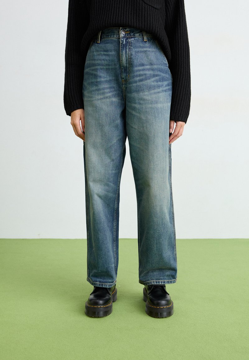 Jeans azzurri a gamba larga in denim, con vita alta e sfumature delicate, abbinati a stivaletti neri con zeppa. Sfondo verde pavimentato.