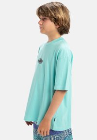 Camiseta de algodón turquesa con cuello redondo y mangas cortas, que presenta un pequeño logo circular en el pecho. La tela tiene una textura suave.