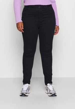 Osoba ubrana w czarne spodnie typu slim fit, białe i szare trampki oraz lawendowy prążkowany top z długim rękawem, stojąca na jednolitym tle.