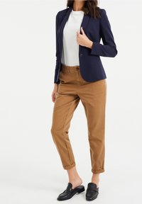 Laivastonsininen bleiseri valkoisen paidan päällä, yhdistetty vaaleanruskeisiin chinssihousuihin ja mustiin liukuisiin kenkiin. Bleiserissä on yksittäinen nappi ja kapeahko leikkaus.