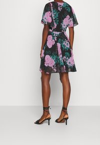 Robe noire à fleurs avec des fleurs roses et teal, manches courtes à volants, taille cintrée et jupe évasée. Portée avec des sandales à talons noires.
