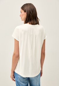 Blouse blanche à manches courtes avec un col rond, détail légèrement froncé au dos et une texture douce et lisse. Portée avec un jean bleu.
