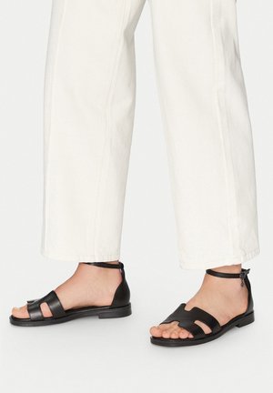 Sandale din piele neagră cu un design cu vârf deschis, având două curele frontale, o curea pe spate cu un cataramă ajustabilă pentru gleznă și o talpă plată.