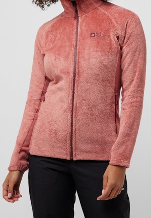 Personne portant une veste polaire rose texturée à fermeture éclair et un pantalon noir, debout devant un fond uni gris clair.