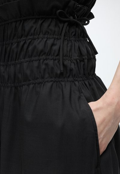 Χέρι που στηρίζεται στην τσέπη μαύρου παντελονιού με ρυθμιζόμενο λαιμόκοψη με κορδόνι στο πάνω μέρος.