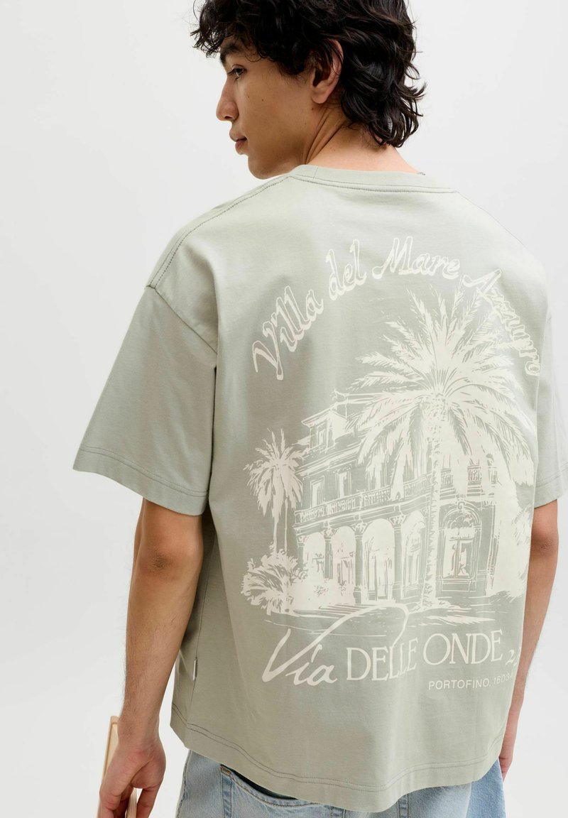 Jeune homme aux cheveux bouclés foncés portant un t-shirt vert pâle avec un graphisme blanc représentant une villa et des palmiers avec un texte en italien.