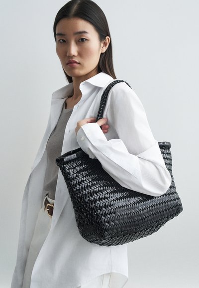 Melna austa iepirkumu soma, taisnstūra forma, ar teksturētu virsmu, aprīkota ar diviem rokturiem. Modelis valkā baltu krekli un pelēku topu, turot somu.