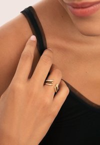 Anello color oro con più fasce, caratterizzato da file alternate di pietre trasparenti e colorate. Mano mostrata su uno sfondo di tessuto scuro.