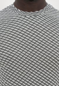 Juodai balta T-shirt su tekstūriniu geometriniu dizainu, nuo kurio priklauso apvalūs motyvai, pasikartojantys tinklelio raštu. Apvalus kaklas.