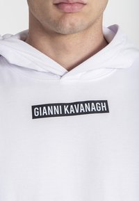 Biely mikina s kapucňou, ktorá má na prednej strane čiernu etiketu s bielym textom "GIANNI KAVANAGH." Tkanina vyzerá mäkko a sú viditeľné detaily šitia.