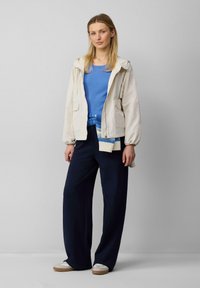 Lichte beige jas met een capuchon, blauwe geribbelde top en losse donkere broek. De outfit bevat witte schoenen met beige zolen.