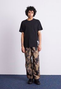 Black short-sleeve t-shirt features a logo on the side. The loose-fit, tie-dye style pants are beige with dark brown and black patterns. Black shoes.