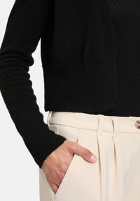 Sweatshirt noir en maille avec des manches côtelées, porté sur un pantalon plissé beige clair, main reposant dans la poche, mettant en valeur la texture et les détails de conception.