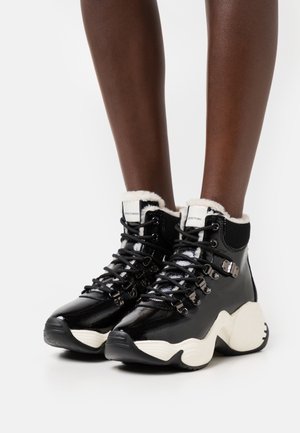Czarne trampki typu high-top z lakierowanej skóry, z białymi masywnymi podeszwami i podszyciem z polaru, noszone na ciemnoskórych nogach na białym tle.