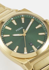 Reloj de metal dorado con un dial verde texturizado, marcadores de hora rectangulares y una ventana de fecha a las 6 en punto. Presenta una correa de acero inoxidable.