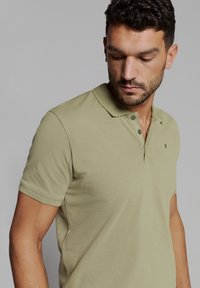 Olijfgroene polo shirt van ademende stof, met een kraag, knoopsluiting met drie knopen en subtiele logo-detail op de borst.