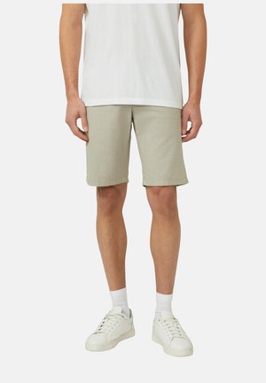 Mężczyzna ubrany w beżowe szorty, białe skarpetki, białe trampki i białą koszulkę stoi na tle jednolitego tła.