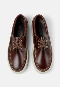 Braune Lederbootsschuhe mit weißen Gummisohlen, heller Naht und Schnürdesign. Mit eingeprägtem Logo auf der Innensohle.
