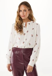 Vrouw met lang golfend haar, gekleed in een witte blouse met kleine bladvormen en hoogwaisted bordeauxrode leren broeken, poserend tegen een witte achtergrond.