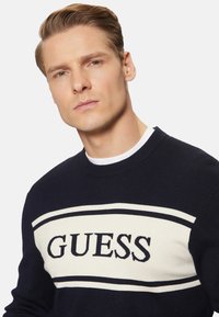 Suéter azul marino con una franja crema en contraste y el logo de "GUESS" en azul oscuro. Presenta un cuello redondo y puños acanalados. Material de punto suave.