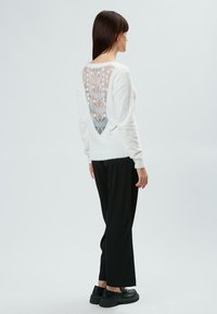 Maglione bianco con un profondo design a V con pizzo sul retro, caratterizzato da motivi floreali, abbinato a pantaloni neri larghi e scarpe nere spesse.