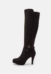 Botas negras de ante sobre la rodilla, con un alto tacón de aguja, tira en el tobillo con un detalle de anillo dorado y una textura suave.
