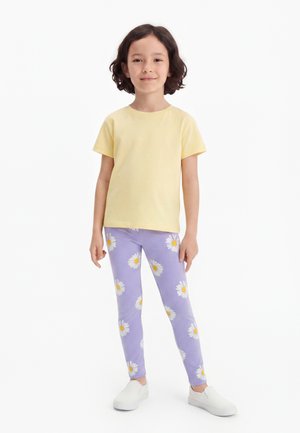 Παιδί με κοντά σκούρα μαλλιά που φοράει κίτρινη κοντομάνικη μπλούζα, μωβ κολάν με λευκά μαργαρίτα λουλούδια και λευκά παπούτσια τύπου slip-on, στέκεται με αυτοπεποίθηση.