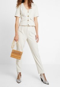 Femme portant un blazer crème à manches courtes et un pantalon, tenant un sac à main en bois avec une anse perlée, chaussant des bottines à motif peau de serpent.