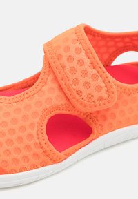 Pomarańczowy but z oddychającej tkaniny z perforowaną teksturą, z rzepem, z wycięciami zapewniającymi wentylację oraz z różową wyściółką z poduszką. Biała podeszwa.