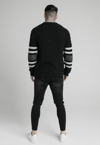 Svart stickad tröja med grå randiga ärmar, parat med svarta skinny jeans och vita sneakers, sedd bakifrån.