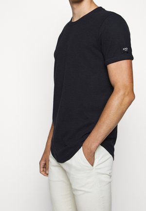 T-shirt noir à col rond en tissu texturé, avec des manches courtes et un patch logo sur la manche. Porté avec un pantalon de couleur claire.