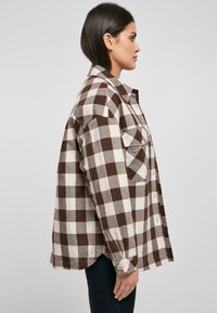 Camisa de cuadros marrón y crema con botones, de manga larga, con bolsillos laterales, un corte relajado y un dobladillo redondeado. Hecha de tela con estampado de cuadros.