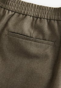Brun, tekstureret stof med elastisk talje og en enkelt baglomme. Har et klassisk design, der er velegnet til afslappet brug.