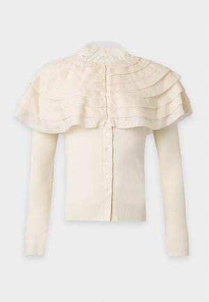 Jonge persoon met een crème kleurige geplooide capelet over een gebreide trui met knopen, brede lichtblauwe jeans en zwarte sandalen met open tenen, staand tegen een witte achtergrond.
