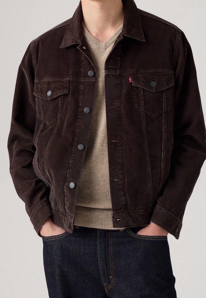Homme portant une veste en velours côtelé marron foncé sur un pull beige à col en V, les mains dans les poches d'un jean en denim foncé.