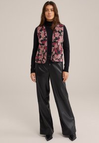 Prsluk s cvjetnim uzorkom u nijansama ružičaste i crne boje, glatke teksture i otvorenog prednjeg dizajna, uparen s crnim hlačama od umjetne kože visokog struka.