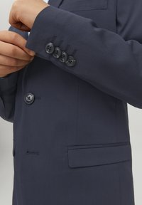 Chaqueta de traje azul marino con textura suave, cuatro botones negros en el puño, cierre de un solo botón y un bolsillo lateral.