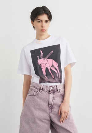 Νεαρό άτομο με κοντά σκούρα μαλλιά, φοράει λευκό T-shirt με ροζ γραφικό γάιδαρου και ψηλόμεσα ανοιχτό μωβ τζιν.