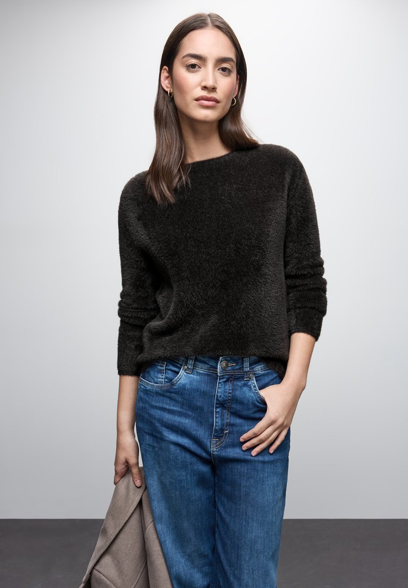 Maglione nero in peluche con maniche lunghe, abbinato a jeans in denim azzurro chiaro leggermente strappati. Cappotto grigio appoggiato sul braccio sinistro.