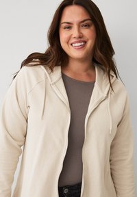 Beige Zip-Hoodie mit strukturiertem Stoff, Kordelzugkapuze und Raglanärmeln, über einem braunen Oberteil getragen. Vermittelt einen lässigen Look.