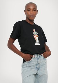 Juodas medvilnės marškinėliai su grafine nuotrauka, vaizduojančia sėdinčią moterį dryžuotu drabužiu, su tekstu "Moschino Jeans" žemiau. Atsitiktinė forma.