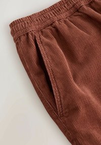 Pantalons en velours côtelé marron avec une taille élastique, des poches latérales et une texture côtelée. Le tissu a un aspect doux et velouté.