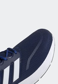 Gros plan sur une chaussure de course bleu marine avec une semelle blanche et trois bandes blanches sur le côté, dotée d’un tissu en mesh et de lacets bleus.