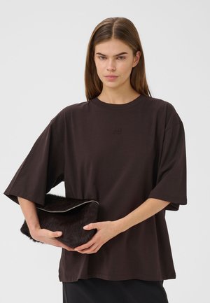 Jeune femme aux cheveux bruns raides, vêtue d'une chemise ample marron foncé, tenant une pochette marron foncé duveteuse devant sa taille.