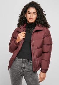 Una donna con capelli ricci che indossa un giubbotto imbottito bordeaux, una maglia nera con stampa leopardata e jeans grigi effetto lavaggio acido, in posa davanti a uno sfondo semplice.