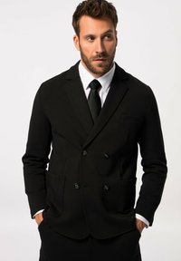 Chaqueta negra de doble botonadura con una textura suave, que presenta solapas en pico, dos bolsillos frontales y detalles en forma de botones. Camisa blanca y corbata negra.