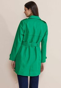 Grön, lätt trenchcoat med knytning i midjan, långa ärmar och axelklaffar. Har en ryggslits för ökad rörelsefrihet.