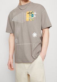 Harmaa puuvillapaita lyhythihaisilla, jossa on geometrinen valkoinen painatus ja keltainen graafinen paikka tekstillä. Yksinkertainen takasuunnittelu.