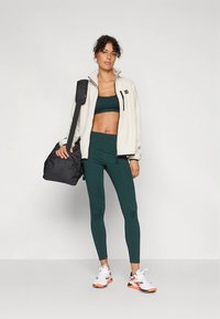 Donna che indossa un reggiseno sportivo verde scuro e leggings, una giacca di pile bianca, scarpe da ginnastica bianche e arancioni, e porta una borsa da palestra nera a tracolla.